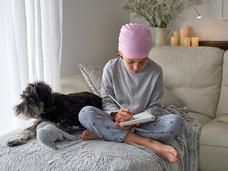 A child wearing a head scarf sits with legs crossed on a sofa, writing in a notebook as a dog sits beside the child.